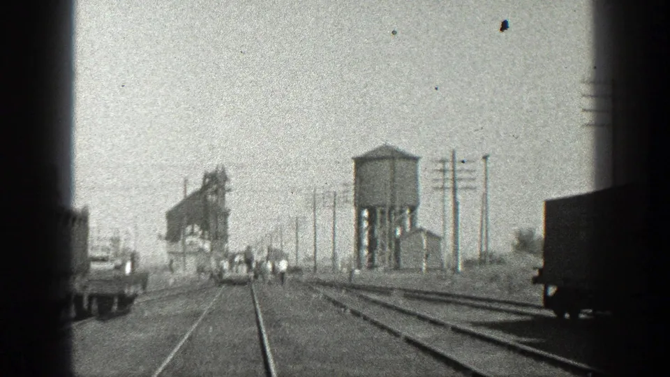 1936 California home-movie frame of an industrial rail yard, showing multiple tracks, a water tower, utility poles, a truck, and distant workers.