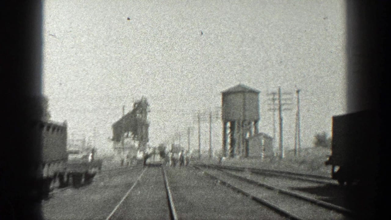 Railroad yard with men walking near tracks, freight cars, utility poles, a water tower, and an industrial structure.
