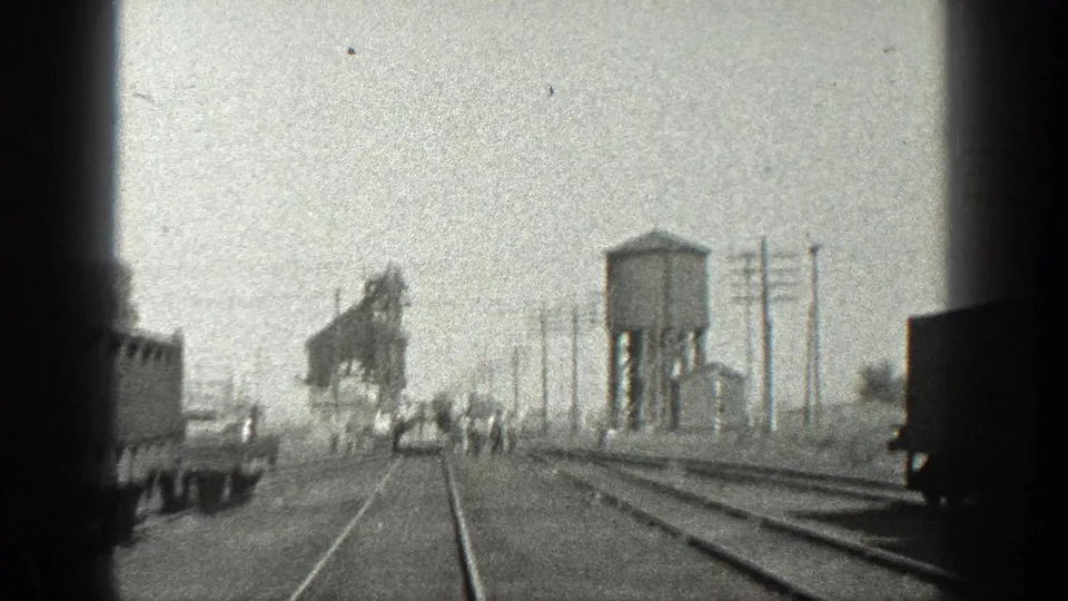1936 California home-movie frame from a moving train showing men crossing a rail yard near freight cars, poles, a water tower, and industrial equipment.