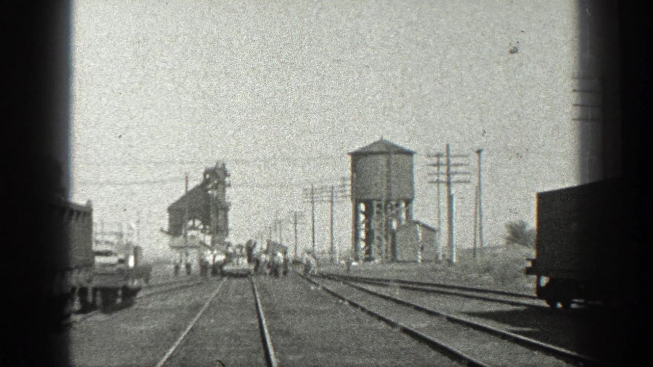 Rail yard view with several men near the tracks, a water tower, utility poles, freight cars, and an industrial structure.