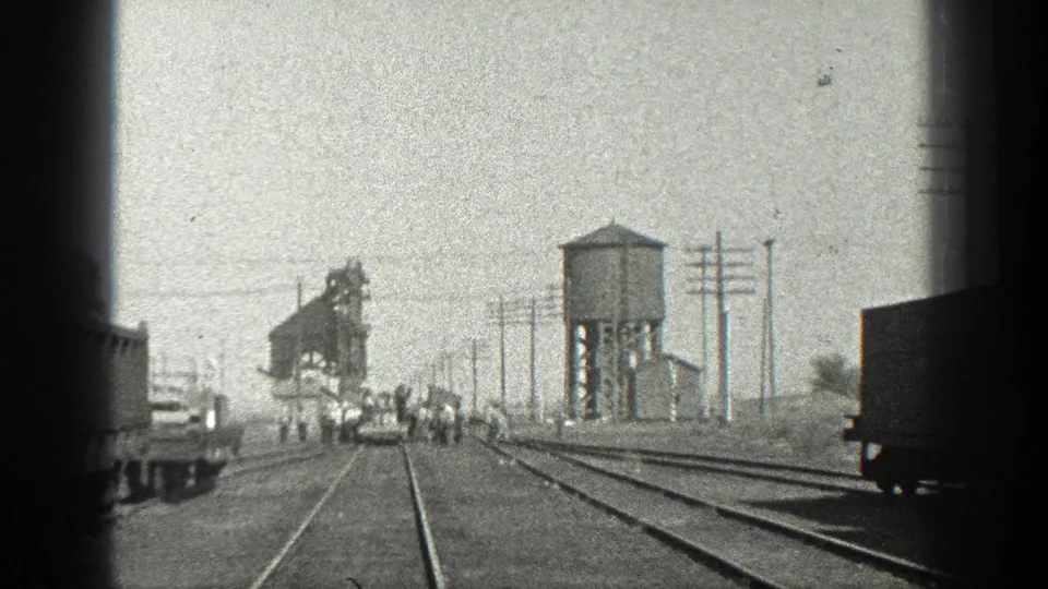 1936 California rail-yard view with small figures walking across tracks near a water tower, freight cars, utility poles, and an industrial structure.