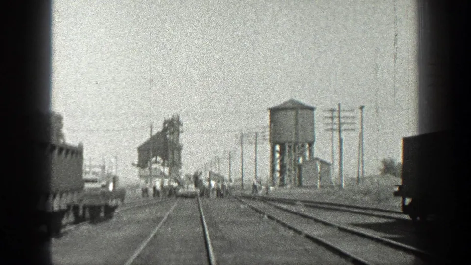 Black-and-white 1936 California frame showing a moving-train perspective on tracks, distant men, freight cars, a water tower, and utility poles.