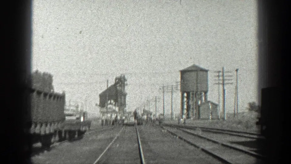 1936 California rail-yard frame with receding tracks, freight cars at the edges, distant men, utility poles, a water tower, and industrial structures.