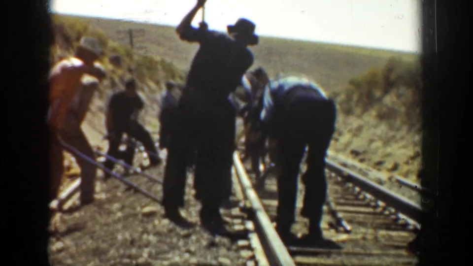 Men maintain railway tracks in a rural California setting, filmed in color on 8mm with visible gate edges and soft focus.