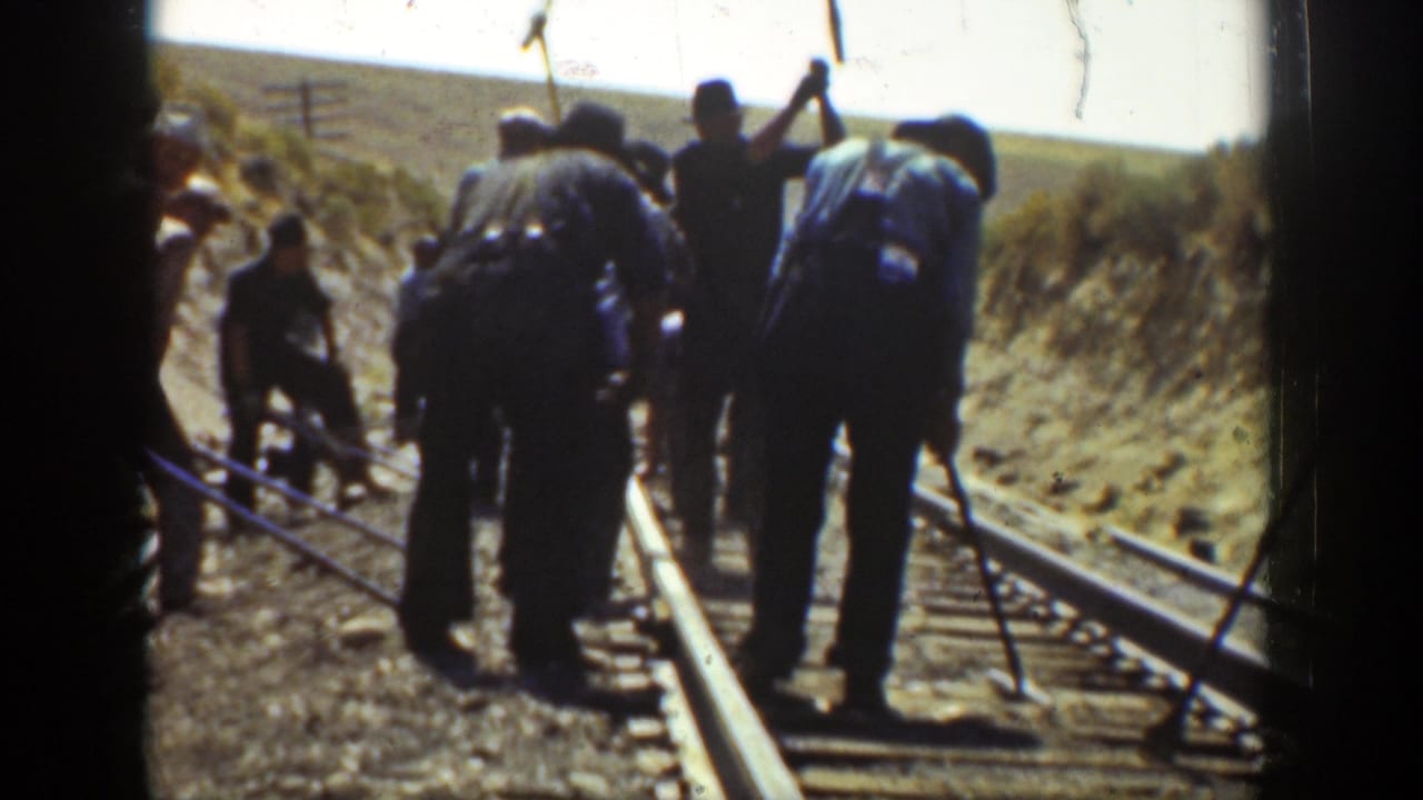 Men in work clothes use tools on a railroad track in a dry rural landscape.