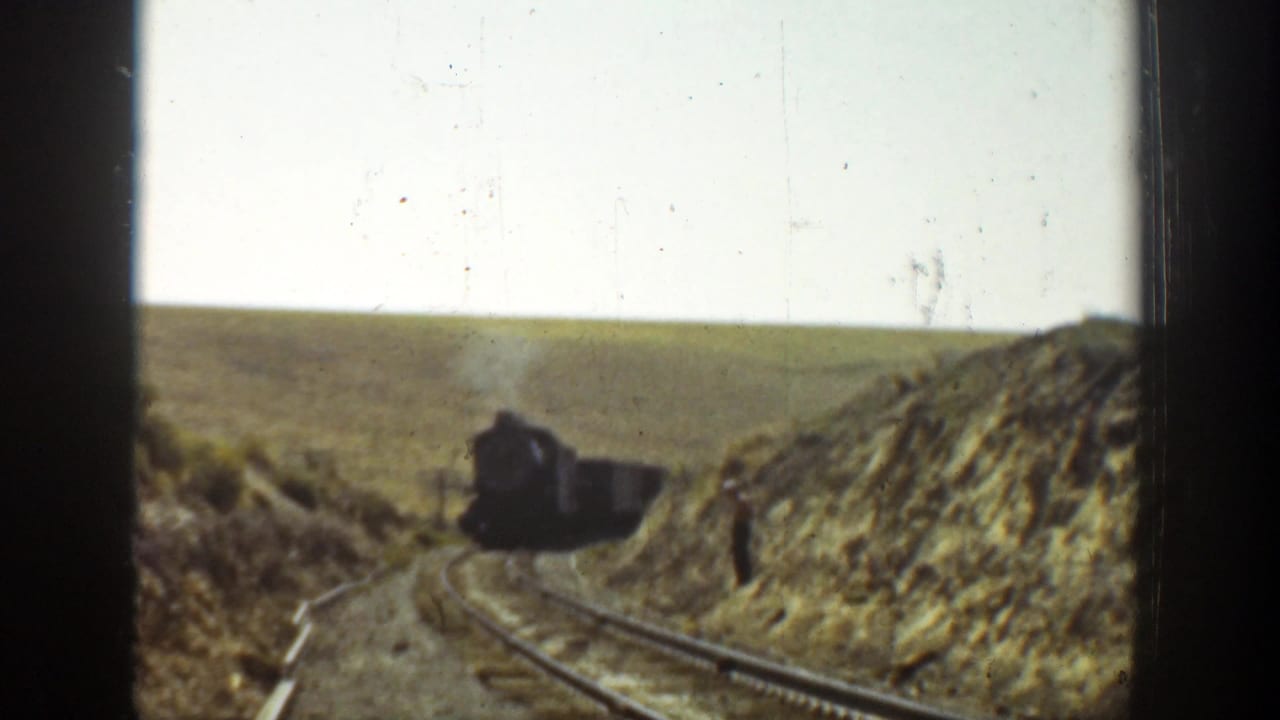 A dark train approaches around a curve through a cut in open hills.