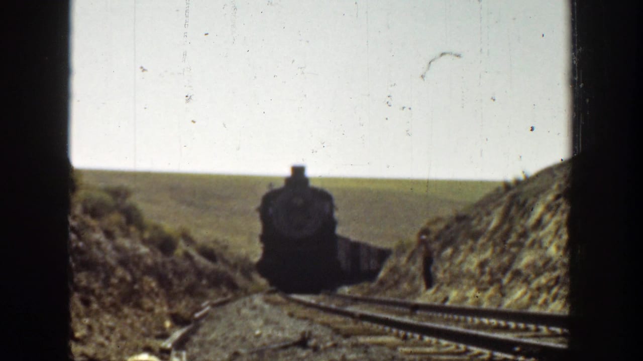 Black steam locomotive approaches on tracks between grassy embankments under a bright sky.