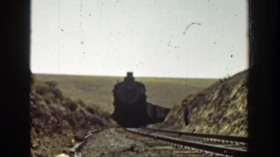 1936 California 8mm frame showing a black steam locomotive approaching through a rural rail cut with hills and sky behind it.