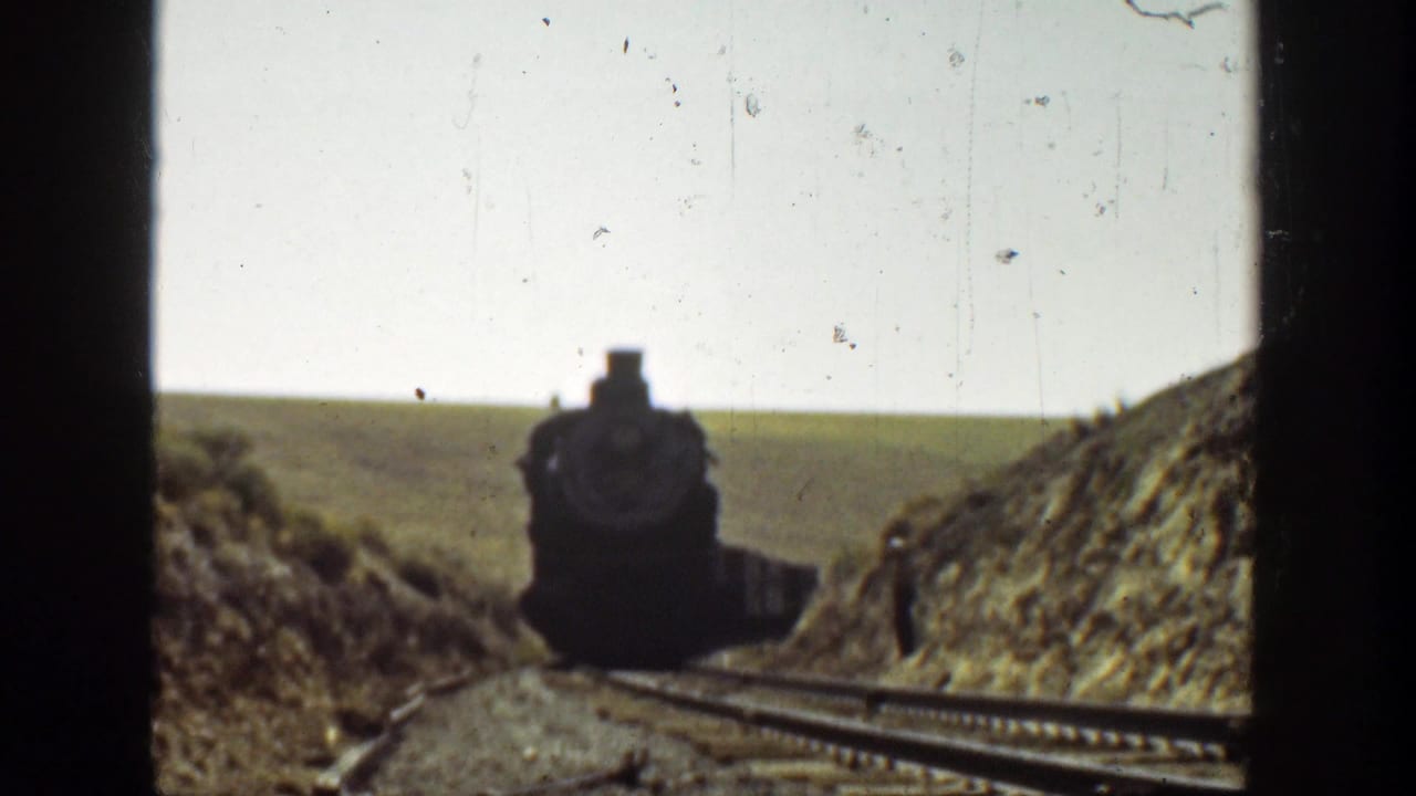 Steam locomotive draws closer on curving tracks with a person standing near the right embankment.
