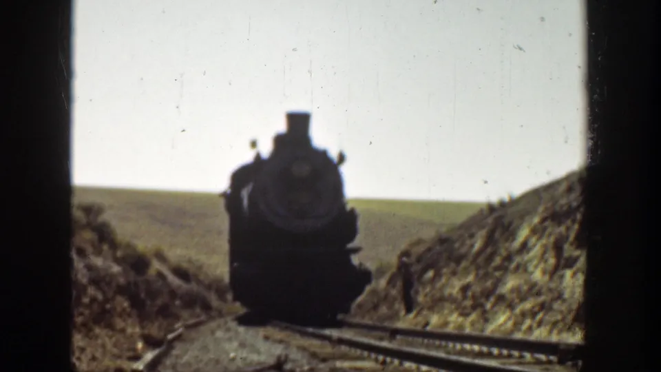 1936 California home-movie frame showing the locomotive close to the camera, with rails, embankment, open hillside, and a small trackside figure.