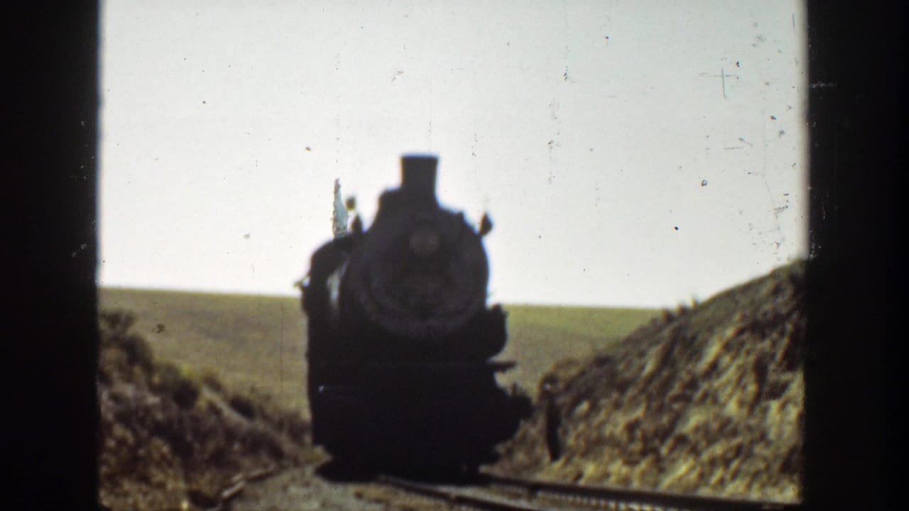 Steam locomotive approaching along tracks between low embankments.
