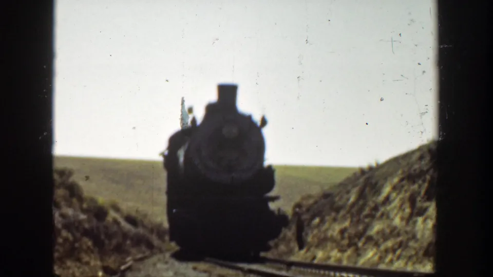 A dark steam locomotive approaches on railroad tracks in 1936 California home-movie footage, framed by dry valley banks and bright sky.