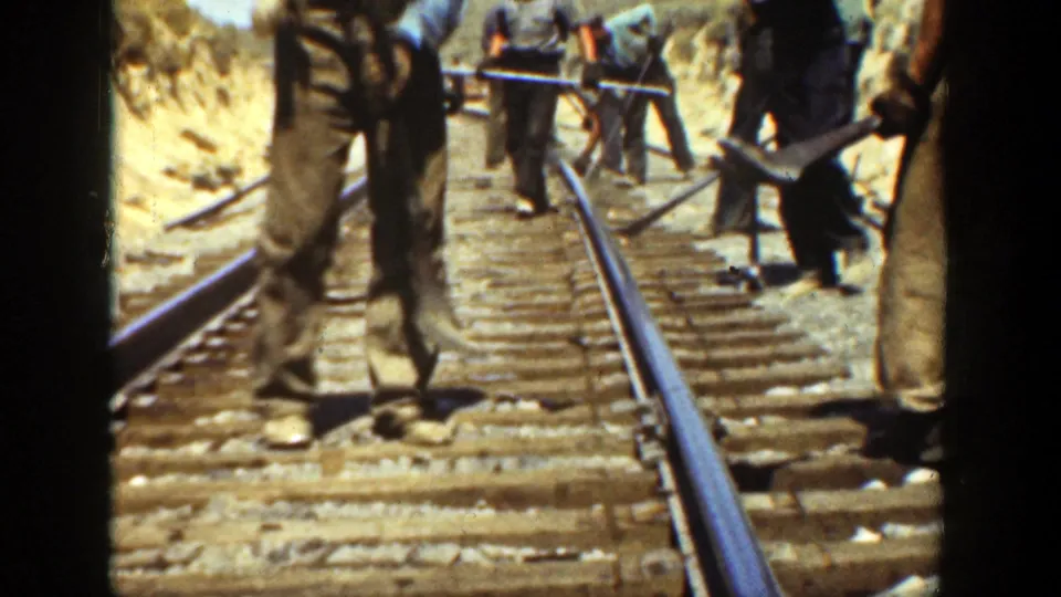 Several workers are visible along the railroad line, using hand tools around the rails and ties in a sunlit California rail cut.