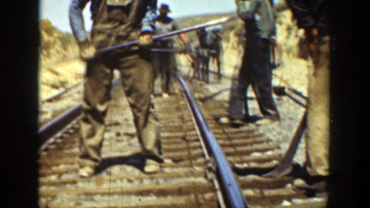 Men stand on railroad tracks with long tools beside rails and wooden ties in a dusty cut.