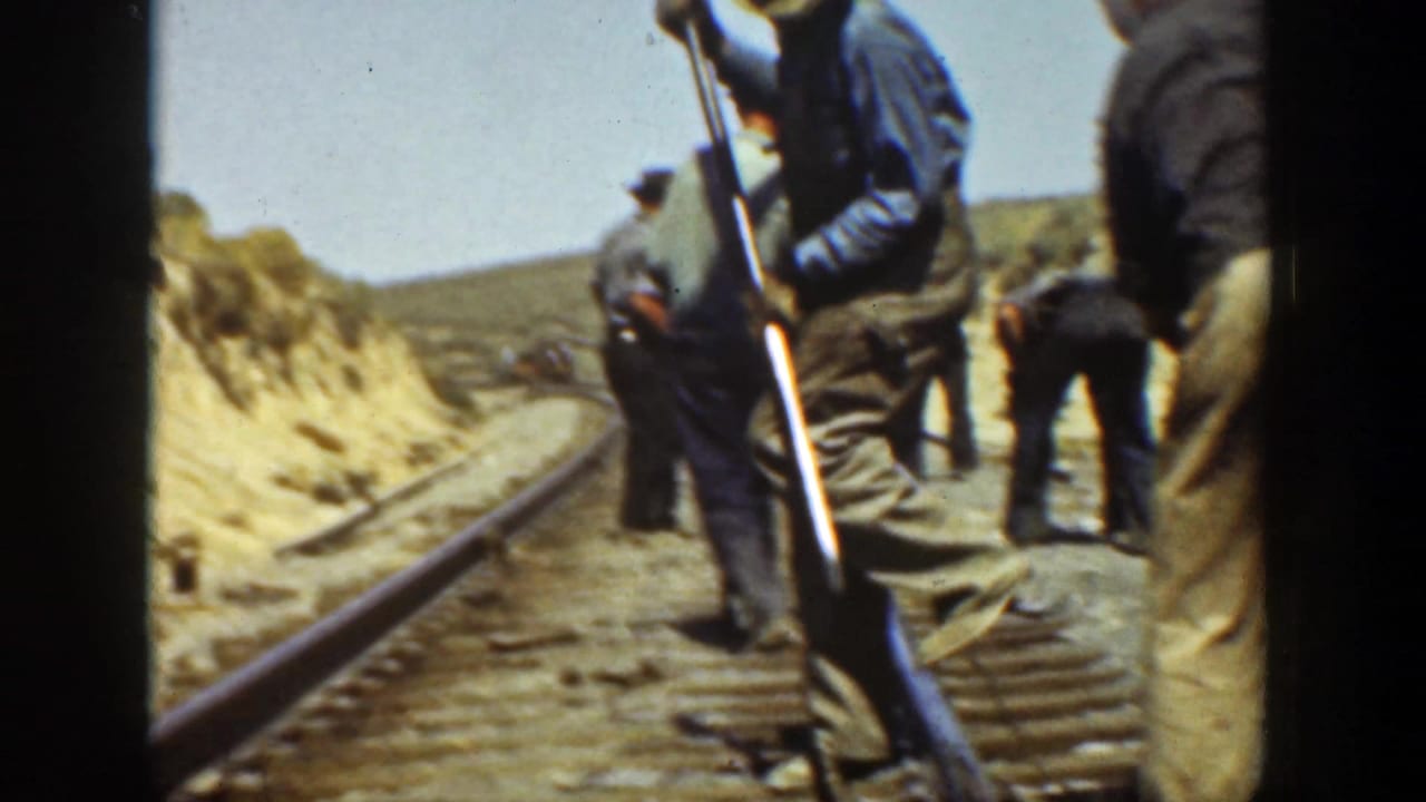 Workers stand and bend near a rail line running through a dusty landscape.