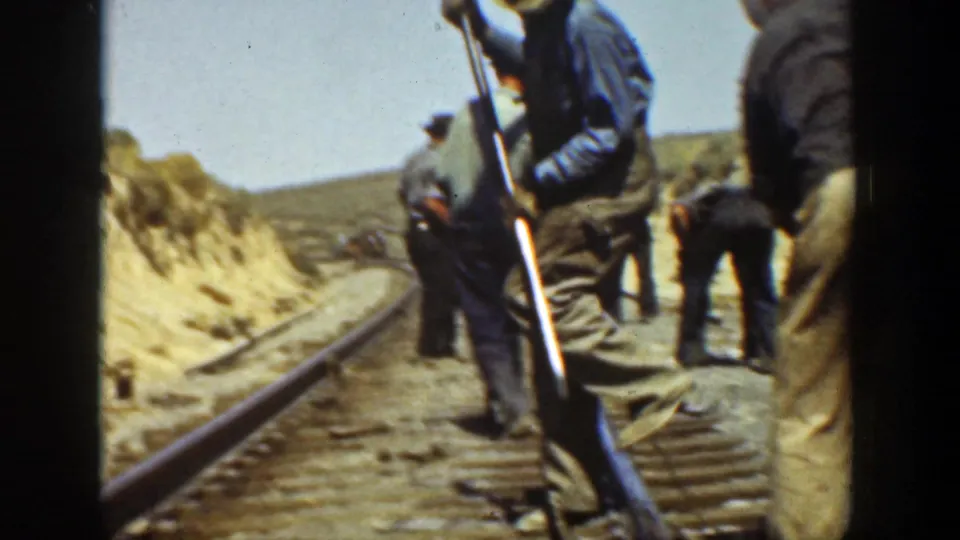 Adult workers in hats and work clothes handle tools along a railroad track in arid California terrain.