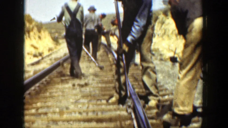 The camera follows adult railroad workers from behind as they move along the ties with tools in a 1936 California setting.