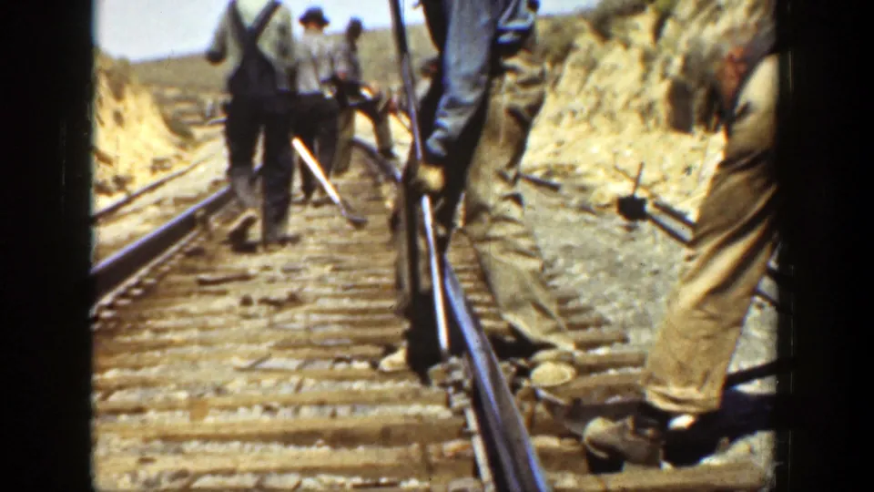Adult workers continue along the rail line with hand tools, framed by dusty banks and distant California hills.