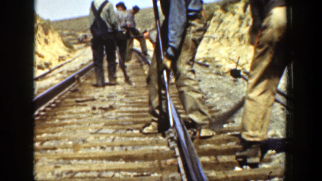 Workers stand and bend over railway tracks beside a dirt embankment on a sunny day.
