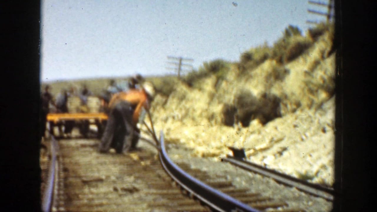 Workers gather near a small cart while repairing curved railway tracks beside a hillside.