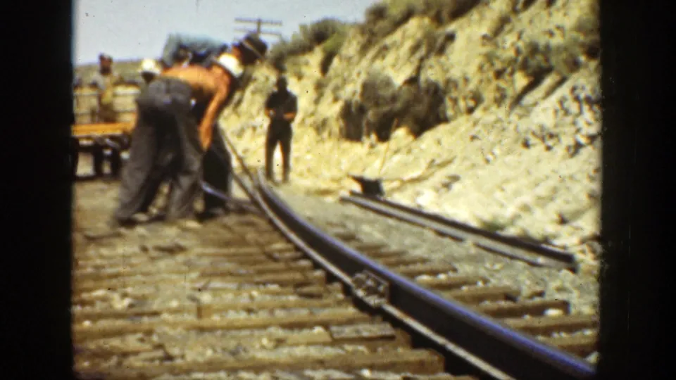 1936 California 8mm frame of railroad workers bending over track with tools, a small rail platform behind them, and a dry hillside in the background.