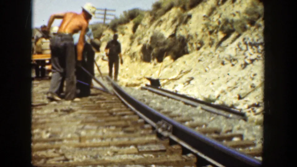 1936 California frame showing a shirtless worker and other men beside railroad rails, with tools, wooden ties, a rocky slope, and utility poles visible.