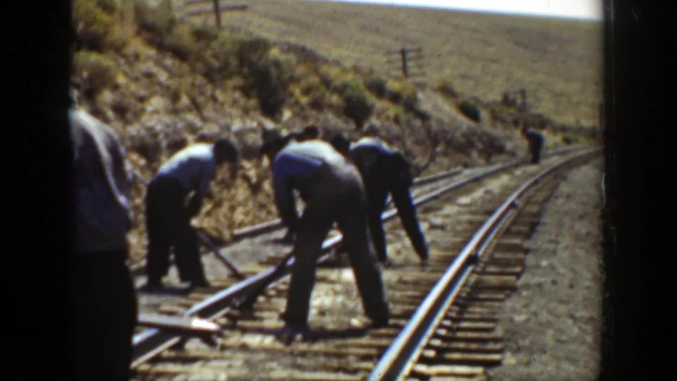 Wide 8mm view of men bending over a curved railroad track in a dry California hillside setting, with one person farther down the line.