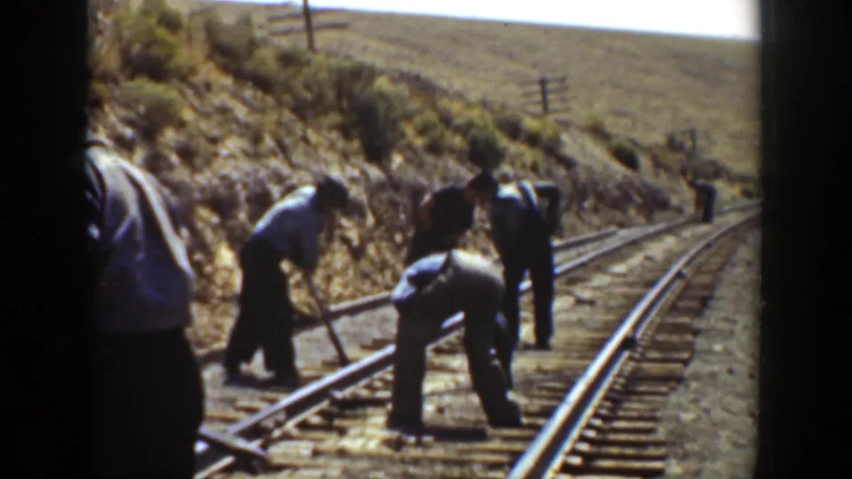1936 California home-movie frame showing workers using tools along curved railroad tracks, with hillside brush and a distant figure visible.