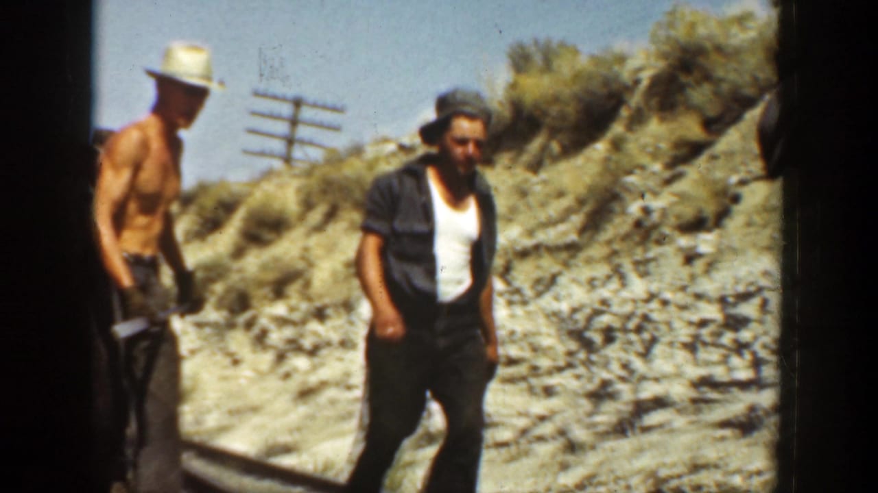 Men stand and work beside railroad tracks in a rocky cut under bright sun.