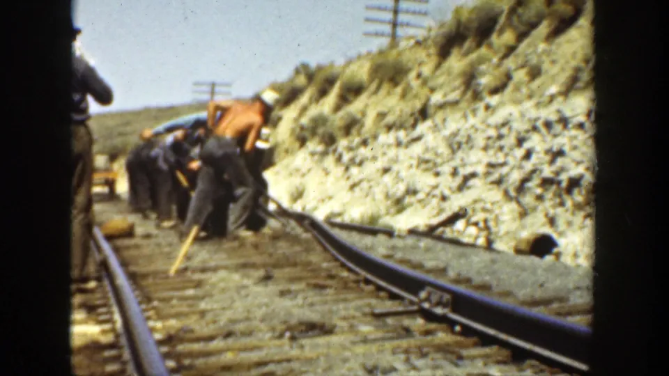 The frame shows men working in a line beside rails and ties, with the camera partly framed by a dark vertical border.