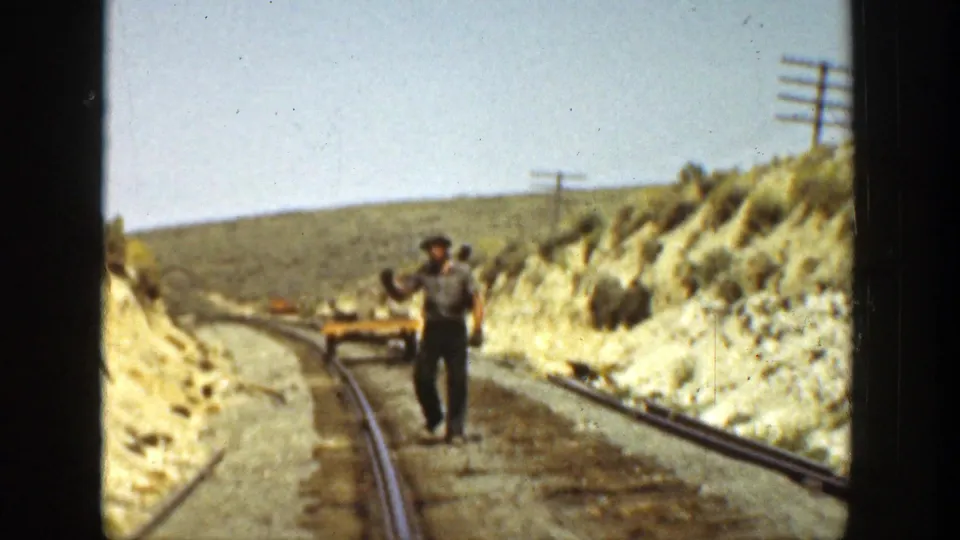 A supervisor-like figure stands on the rail bed in 1936 California footage, with tracks, hillsides, and poles receding behind him.