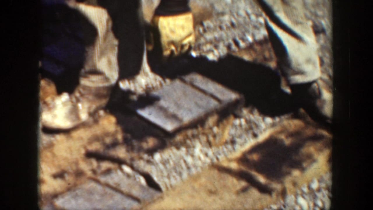 Gloved workers use hand tools around wooden ties and gravel ballast beside a rail.