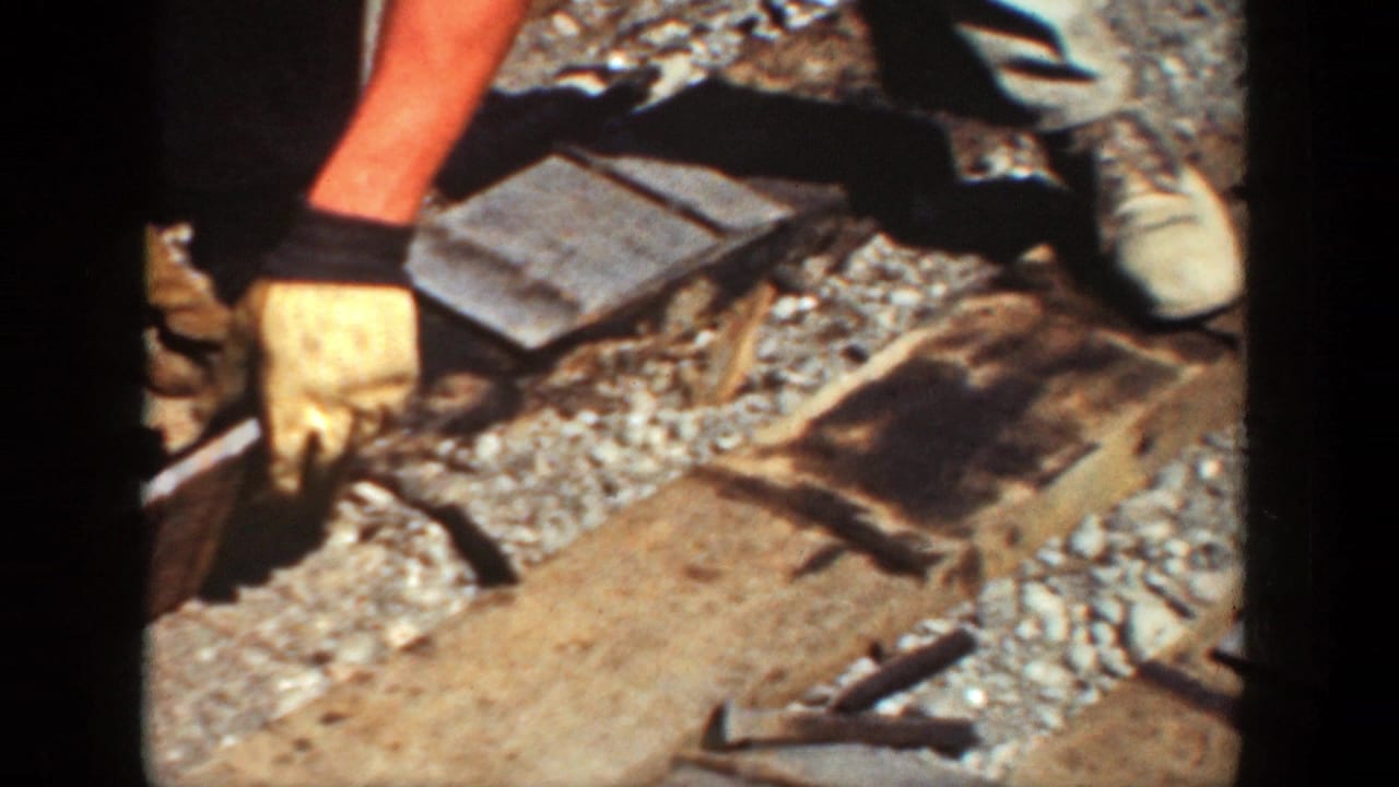 A gloved hand holds a tool near wooden ties, rails, and loose gravel.