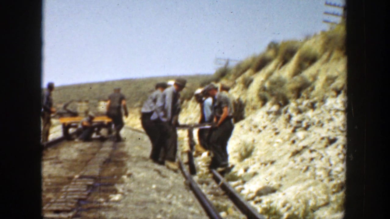 Workers stand and carry equipment along narrow rails in a dry hillside cut.