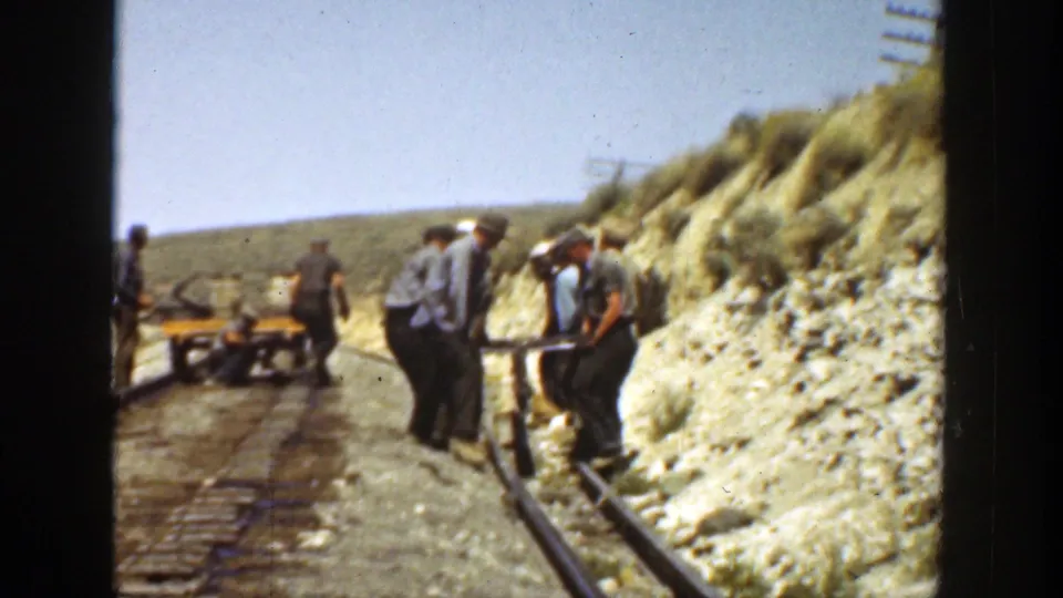 In 1936 California, railway workers appear on a narrow track with equipment and a small handcart in a sunlit, dry hillside setting.