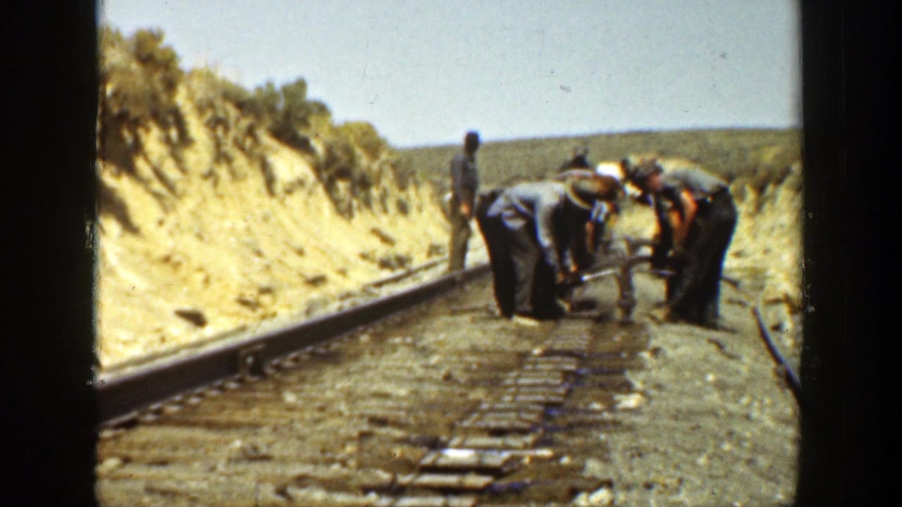 Workers bend over a metal object on railway tracks in a dry landscape.