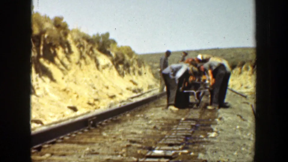 The workers continue bending over the rail bed, coordinating around a heavy object on a 1936 California railway line.