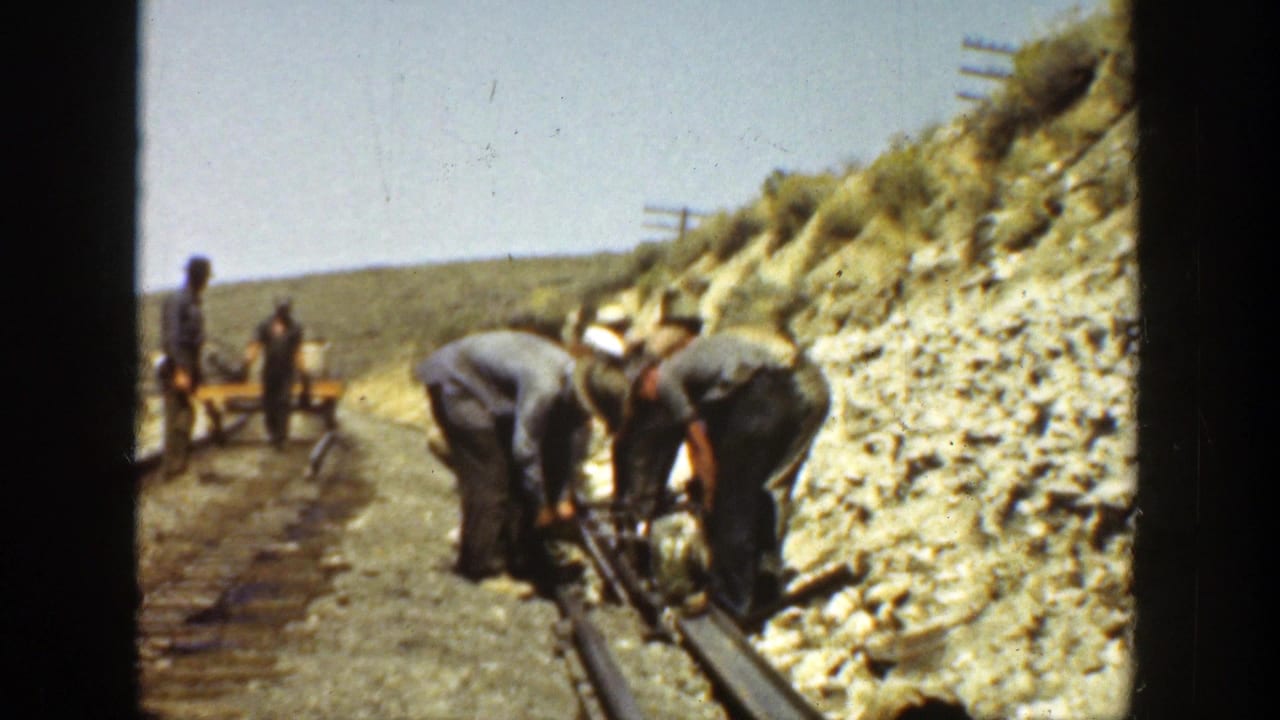 Railroad workers bend over tracks beside a rocky hillside.