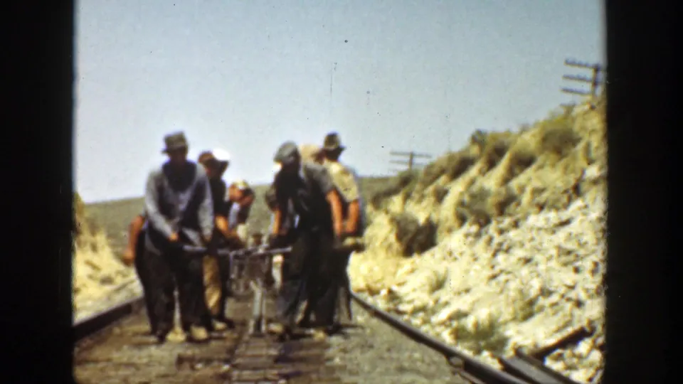 Workers in work clothes and hats guide a small frame or cart on railway track in California archival footage.