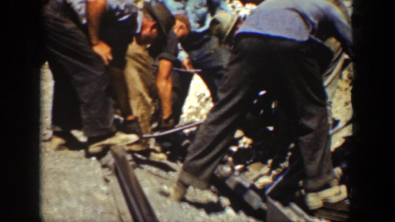 Workers bend over a rail section while handling tools on the track bed.
