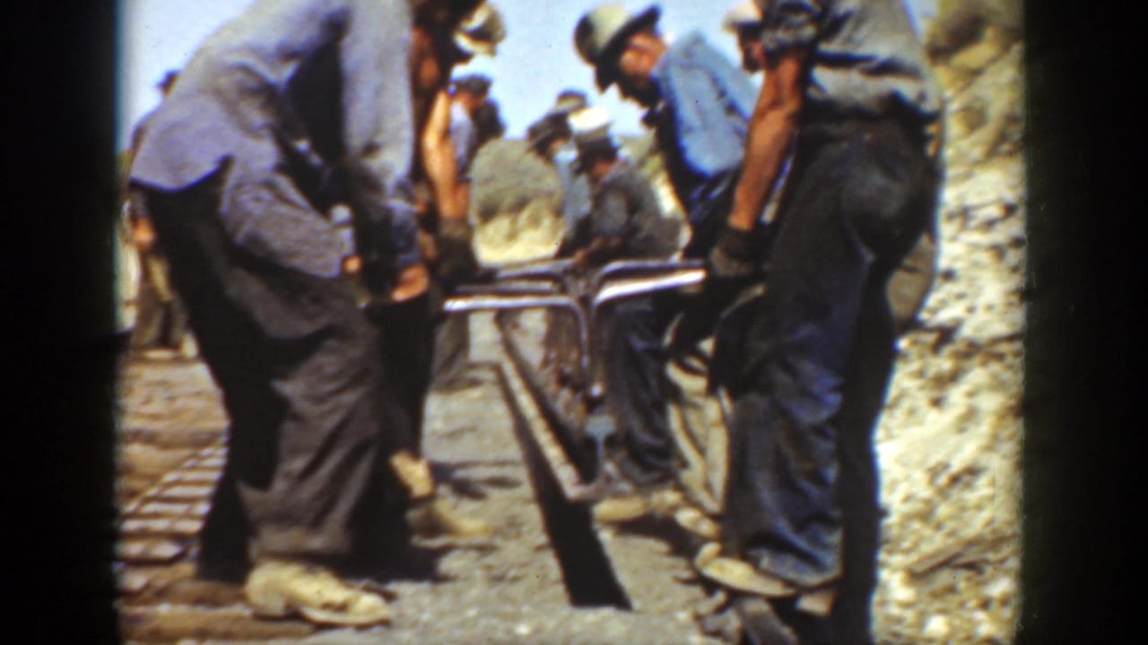 Workers bend over a railway line while handling rail and tools in bright daylight.