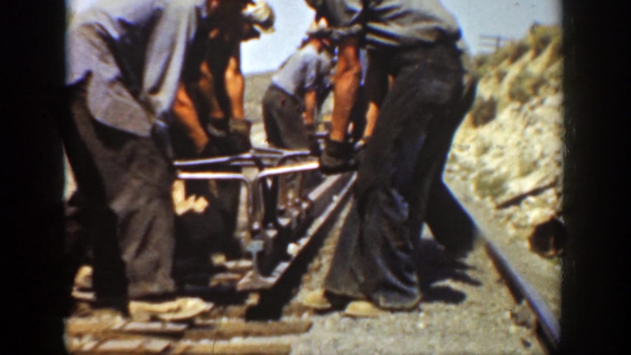 Several workers push or steady a rail tool on tracks beside a rocky bank.