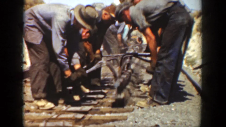 Men in hats and workwear bend over the railway, using hand tools near wooden ties in a 1936 California outdoor work scene.
