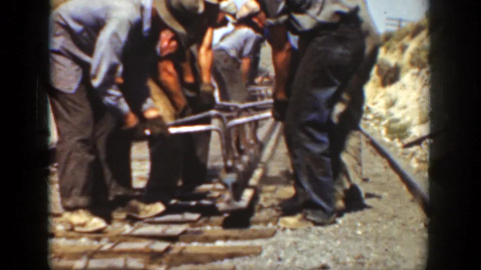 The 8mm frame shows a California rail crew coordinating around a metal rail tool, with iron rails and ties visible under bright sun.