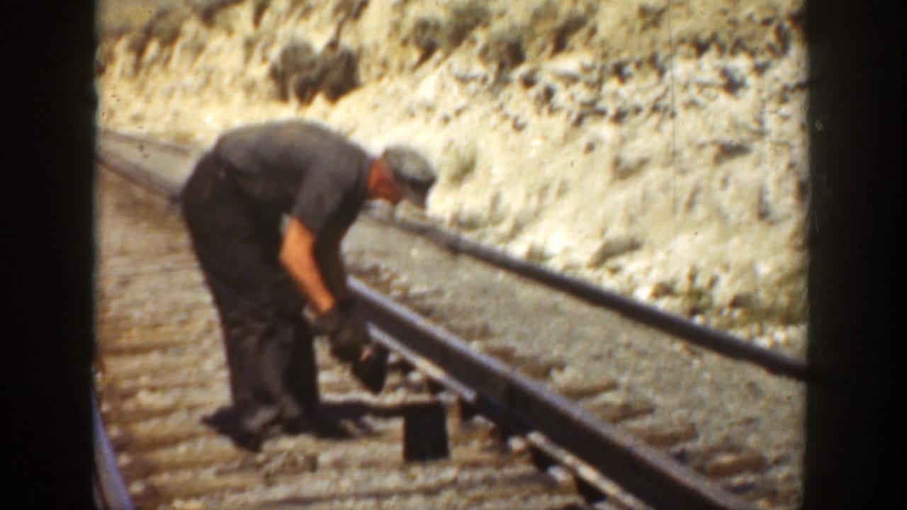 Worker bends over railroad track with a tool beside rails and wooden ties.