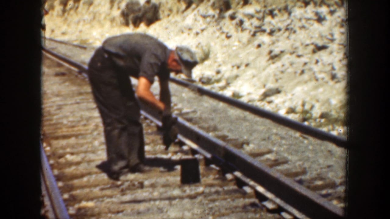 Worker leans over a rail while using a tool on the track bed.