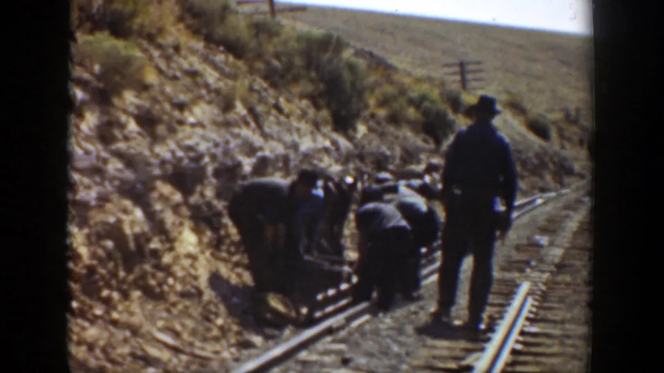 A small railroad crew works low on the tracks while a standing man in a hat observes along a hillside rail line in California.