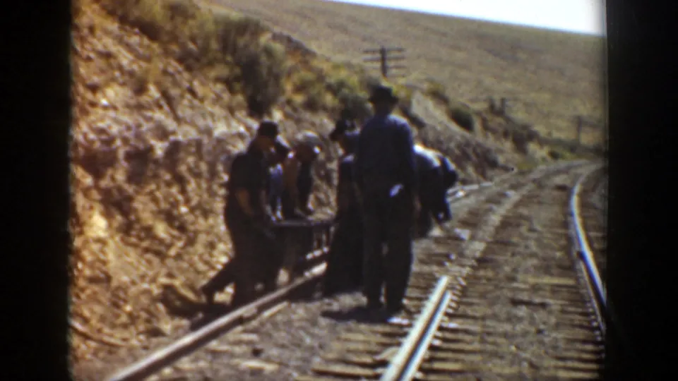 Railroad workers cluster around a small cart or tool carrier on curving tracks, with dry hillsides and utility poles in the background.