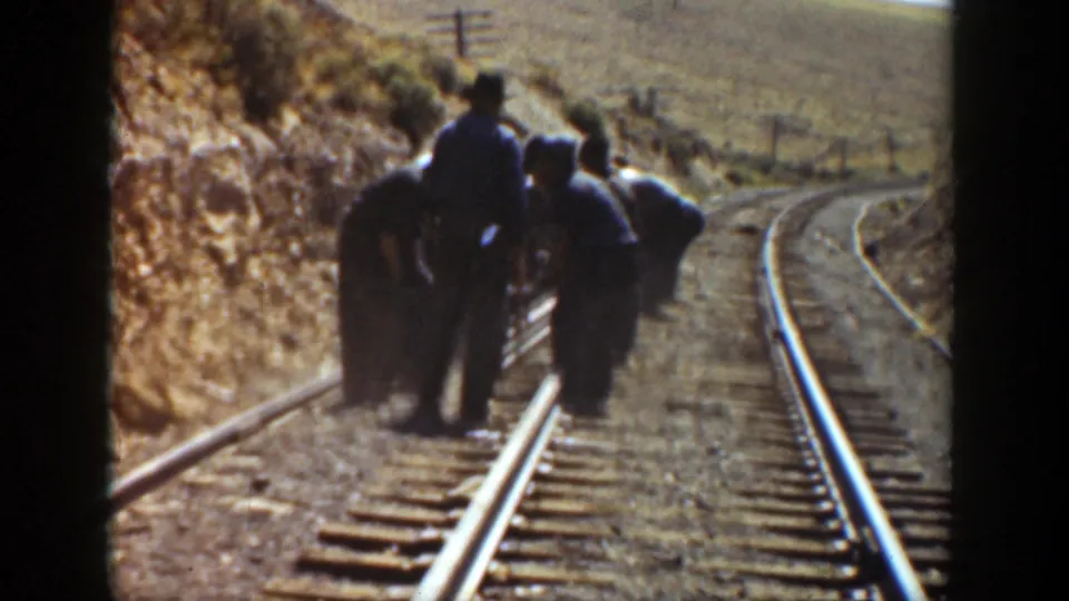 A man in a hat stands beside several men bending near a railroad rail, with dry California hillside terrain behind them.