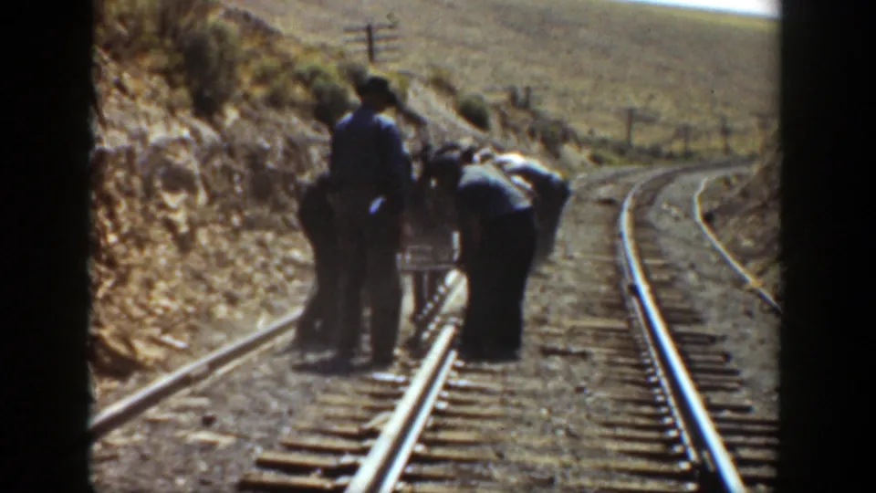 Late frame of 1936 California rail work, with men beside the track and open dry hills visible beyond the rails.
